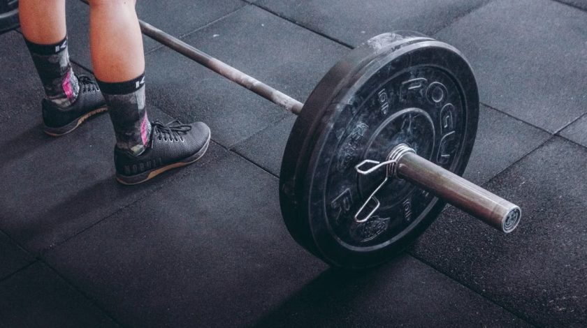 Close-up of an athlete's legs beside a barbell, ready for weightlifting in a gym.